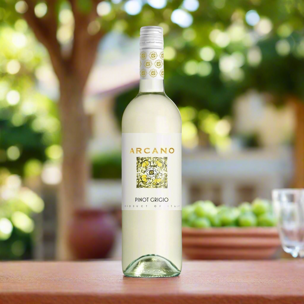 Place a bottle of Arcano Pinot Grigio on a sunlit courtyard table with terracotta tiles, leafy vines, and a carafe of water beside it.