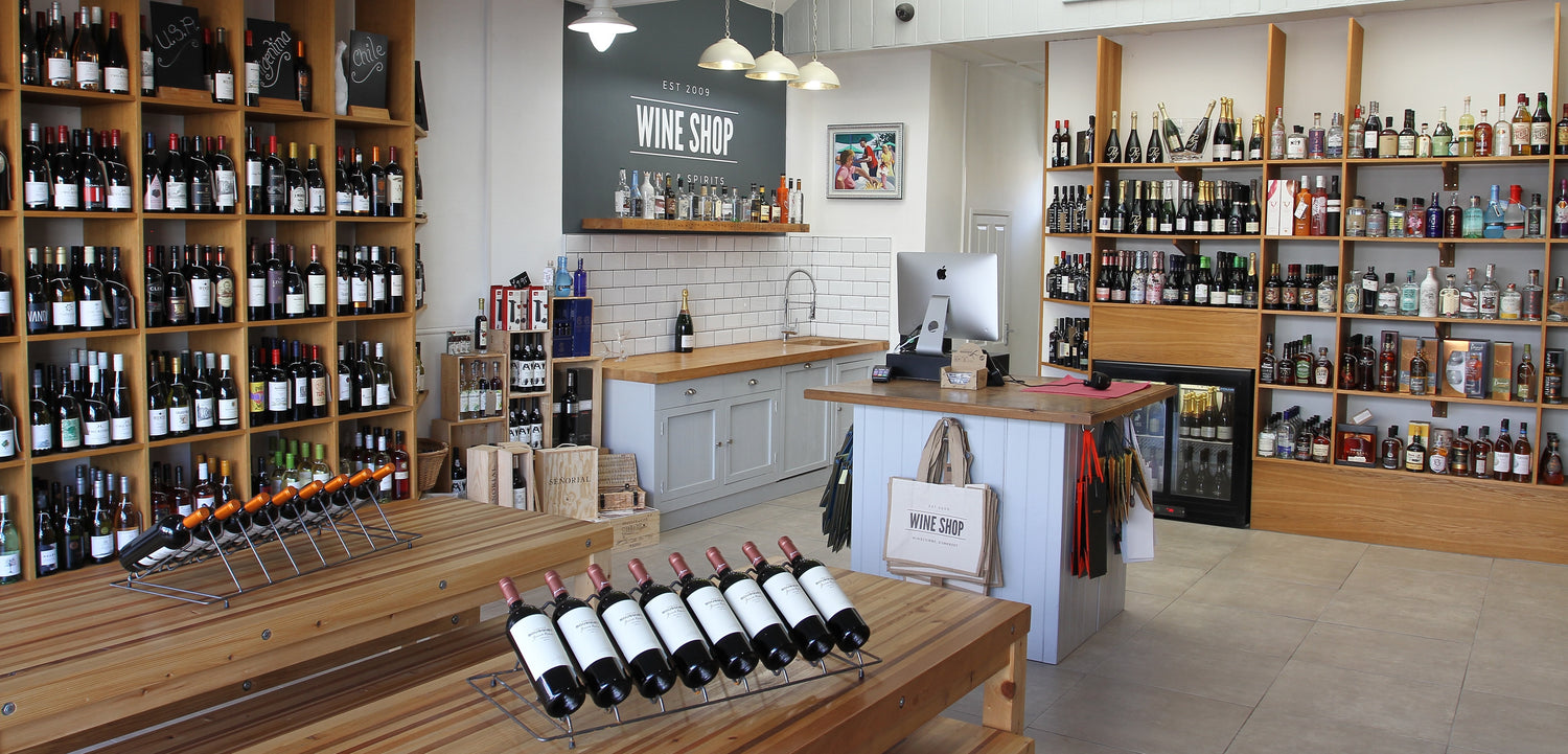 Interior of a modern independent wine shop with wooden shelves, wine bottles, counter top, and branded bags near the till.