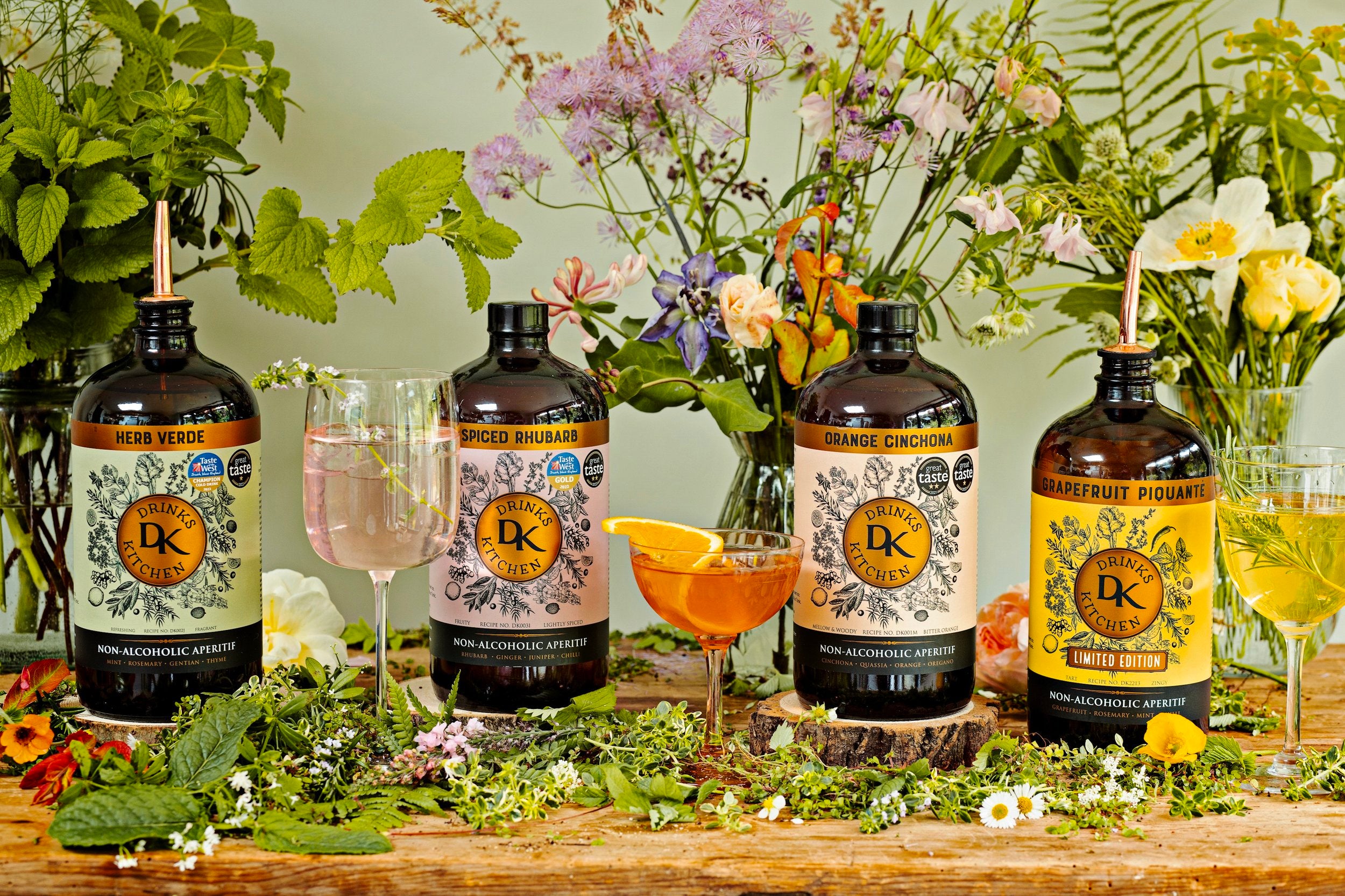Four Drinks Kitchen non-alcoholic aperitifs with garnished glasses on a floral table, surrounded by wildflowers and herbs.