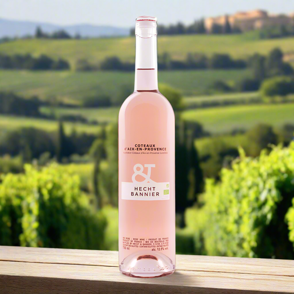 Bottle of Hecht & Bannier Rosé on wooden table in sunlit vineyard with distant Provençal hills.