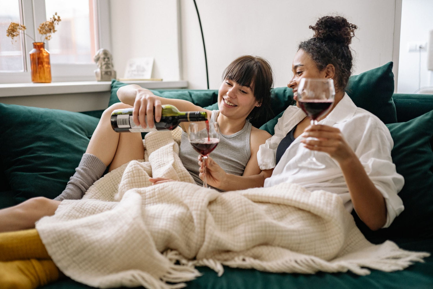 Two friends relaxing on a sofa sharing red wine poured from the bottle on a cosy autumn afternoon