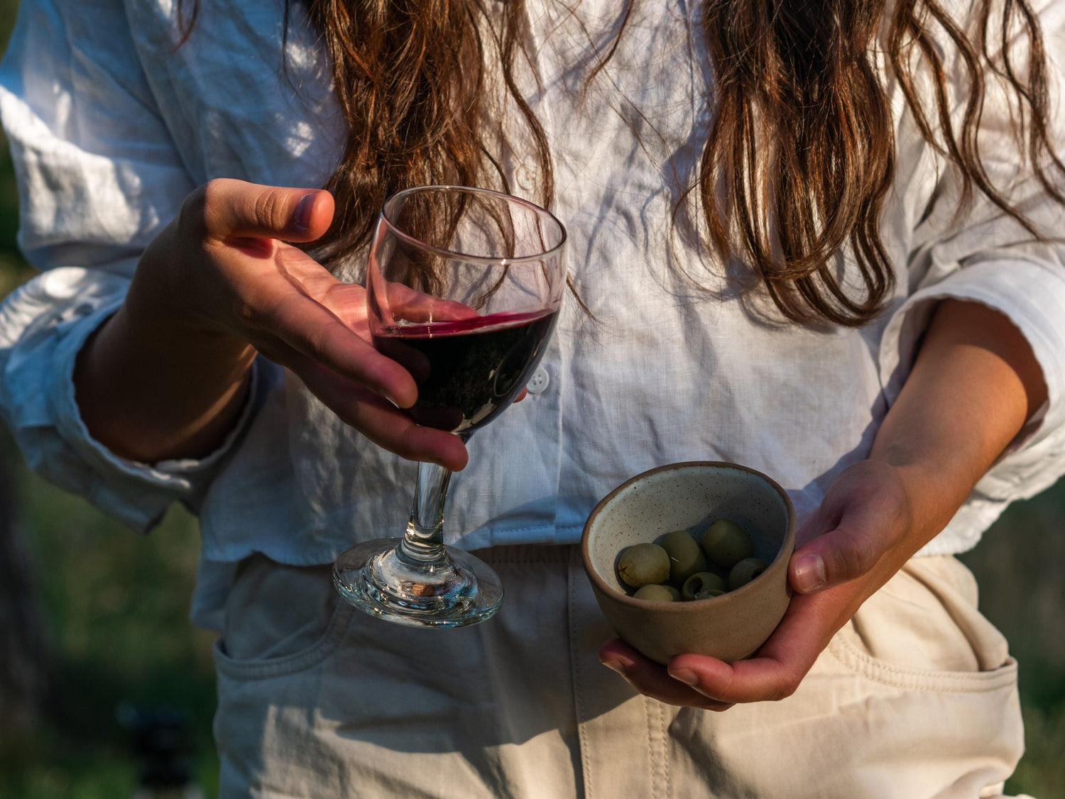 Person holding a glass of red wine and a bowl of green olives in warm afternoon sunlight — Besas Organic