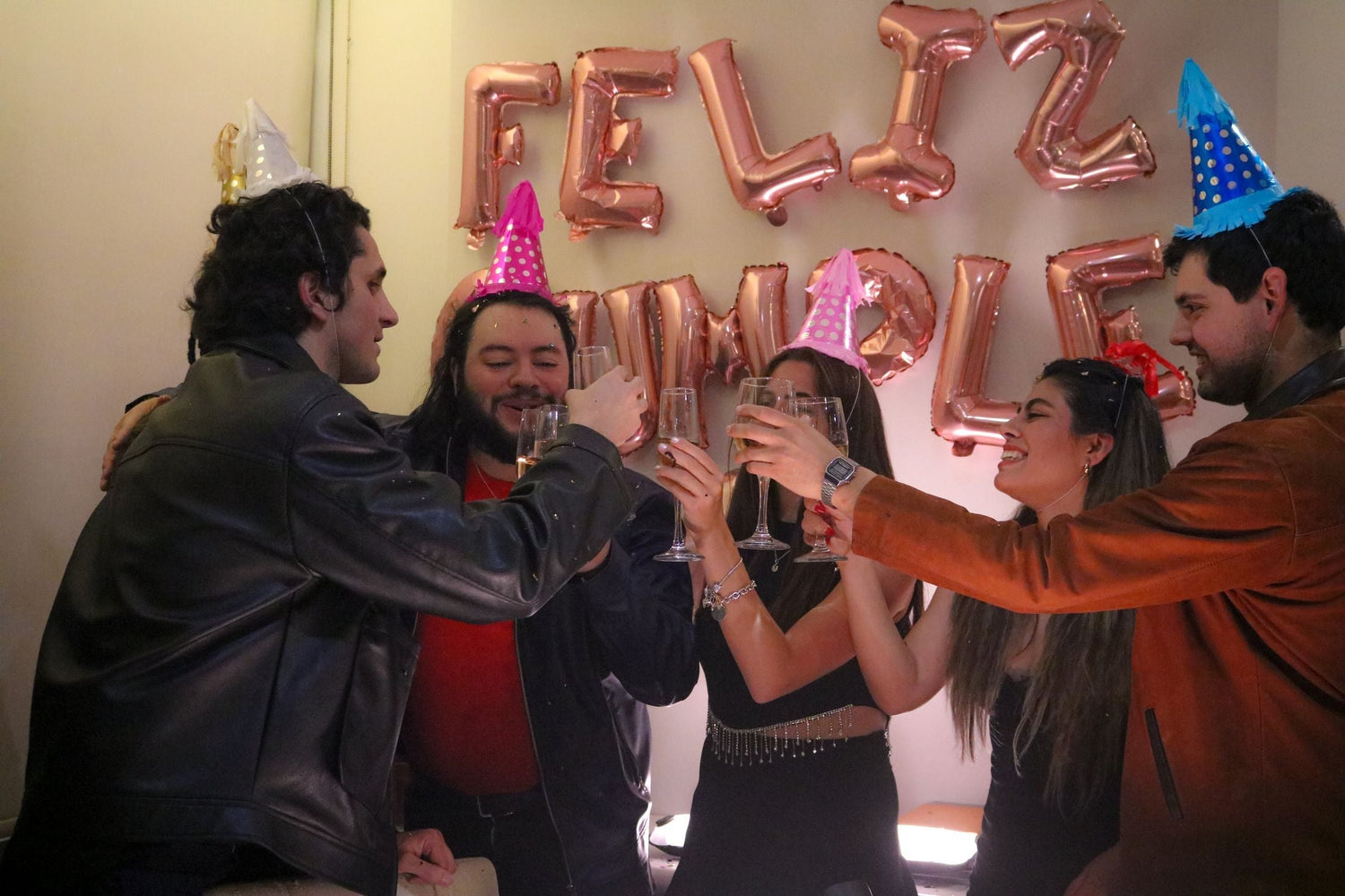 Friends toasting with champagne flutes in party hats beneath birthday balloons — wine birthday gifts