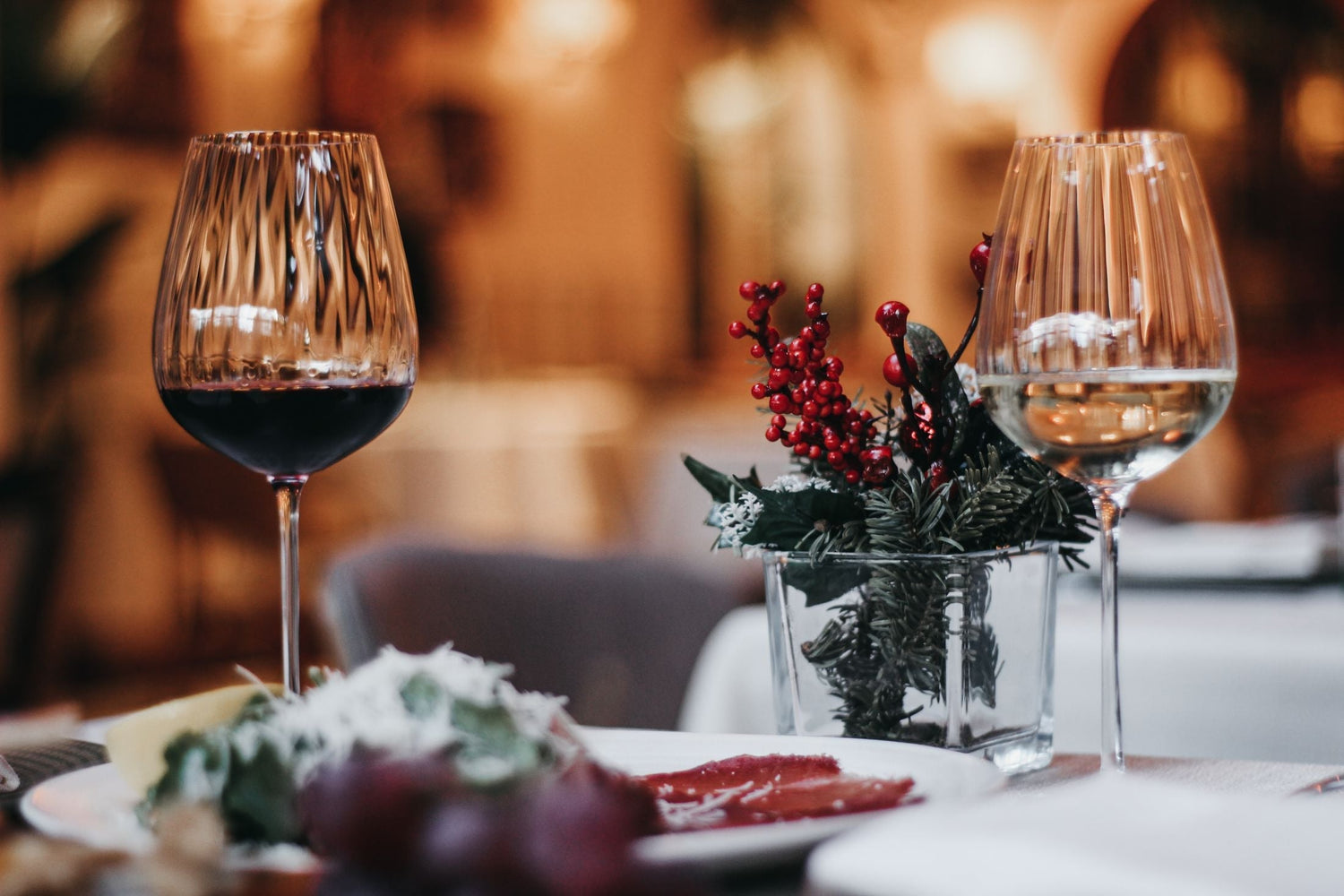 Red and white wine glasses beside a holly and red berry centrepiece on an elegant Christmas dinner table, festive wines for Christmas Day