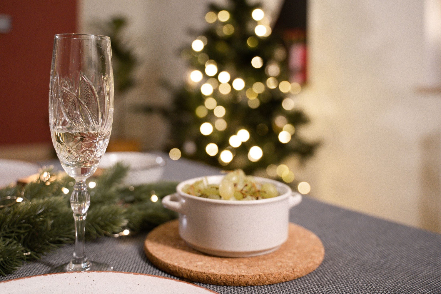 A crystal champagne flute beside a bowl of green grapes with pine sprigs, and a lit Christmas tree glowing softly behind, festive white wine