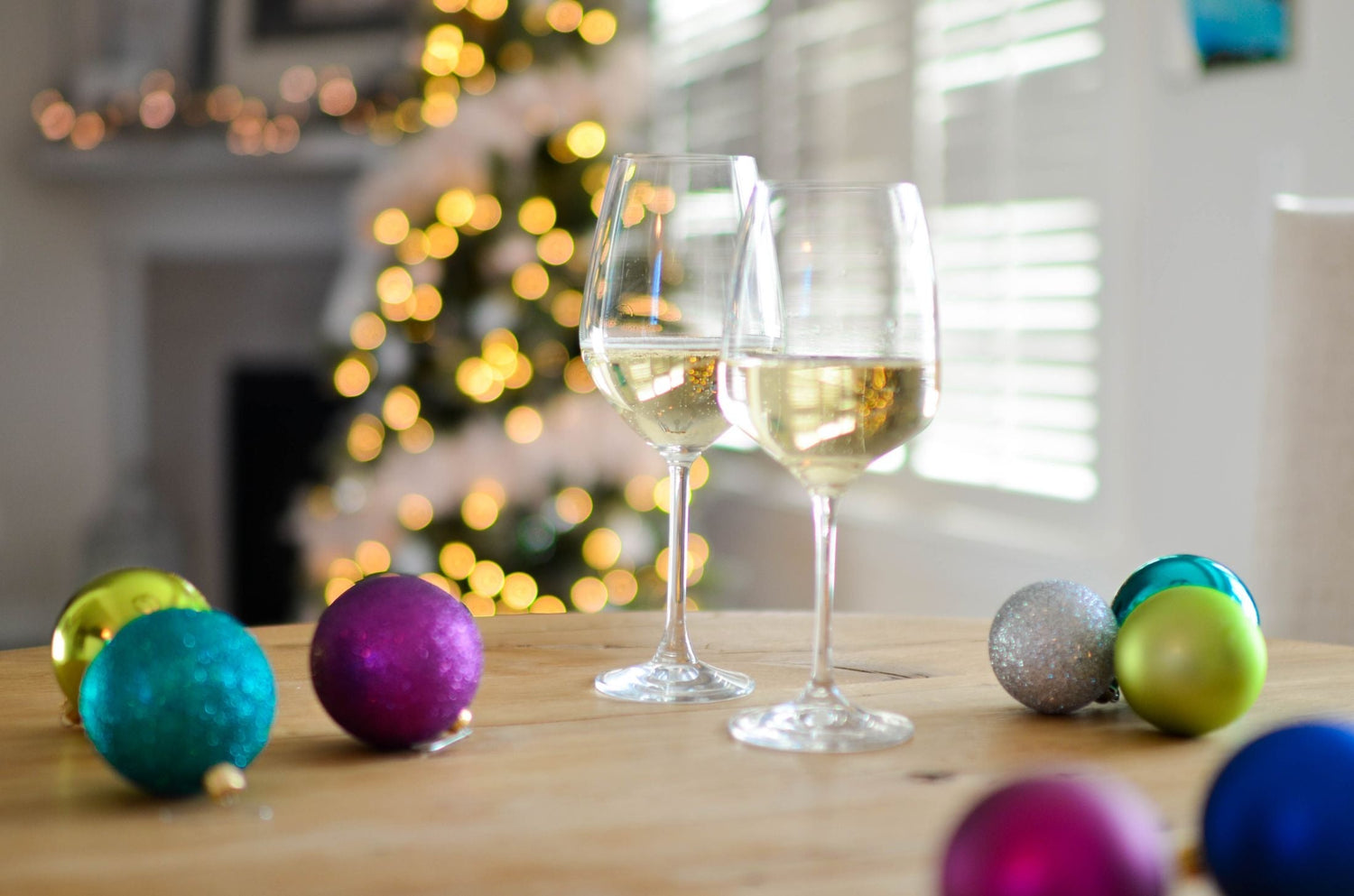 Two glasses of white wine on a wooden table among colourful Christmas baubles, with a lit Christmas tree glowing in the background