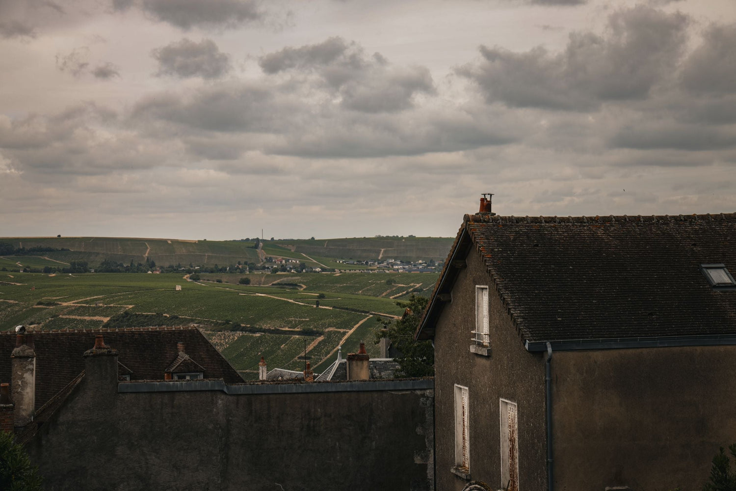 View across slate-roofed village rooftops to rolling vineyard hills under a dramatic cloudy sky, Domaine de l'Amandine, France