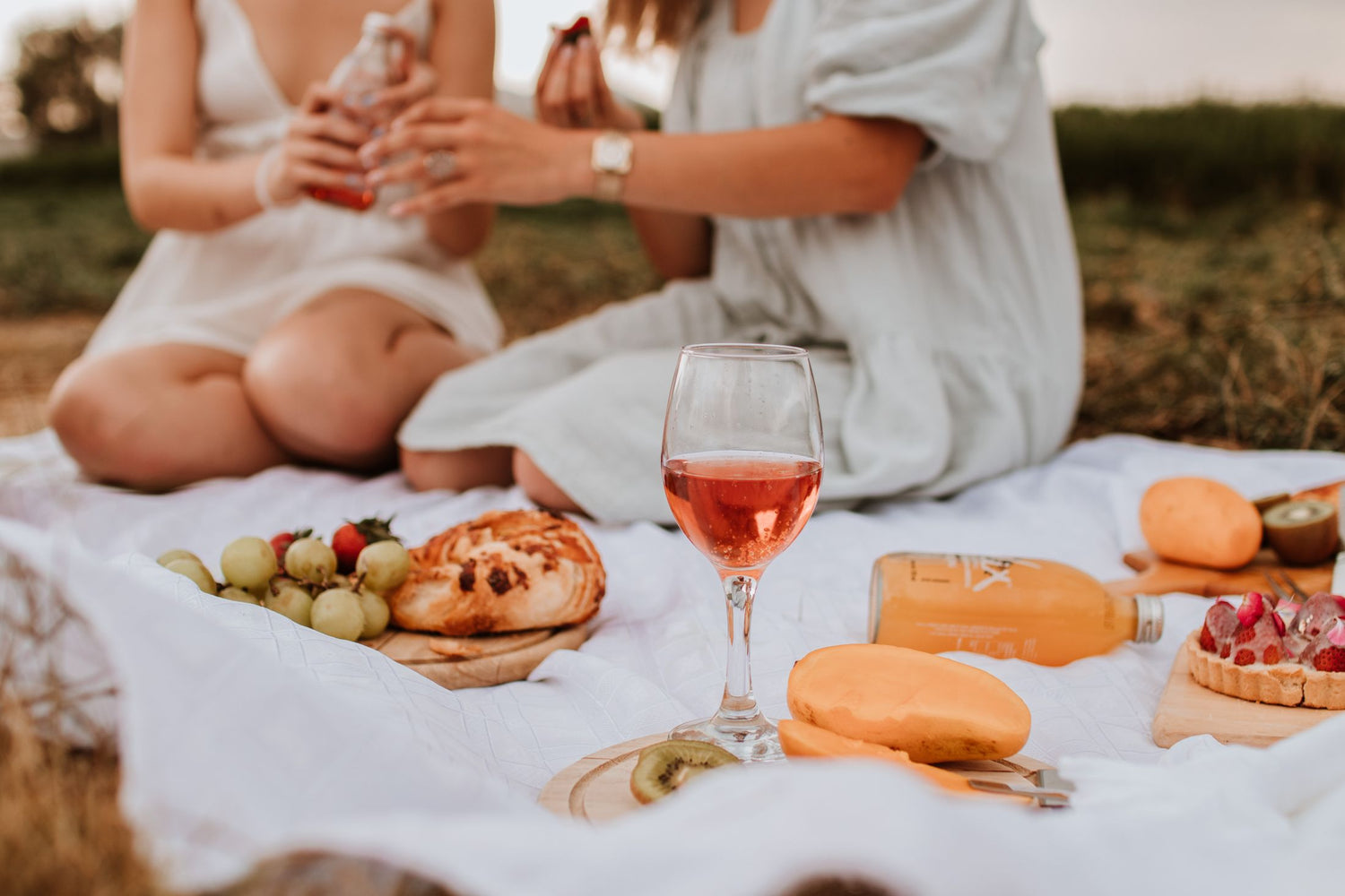 Outdoor picnic scene with a glass of rosé wine, grapes, croissant and fresh fruit on a white blanket on summer grass