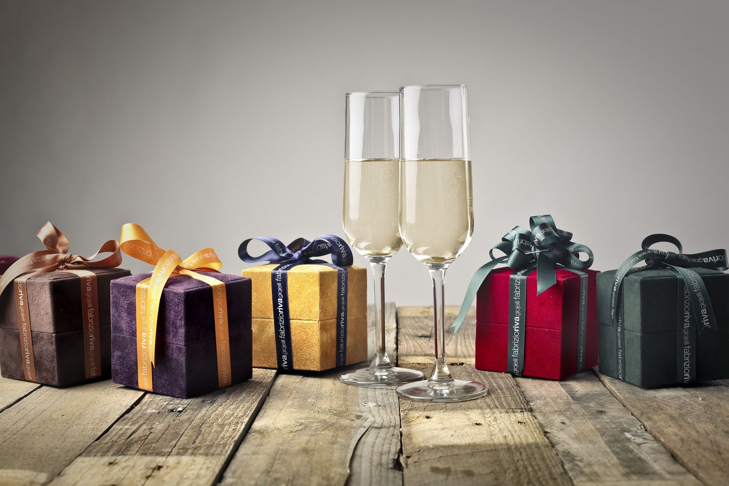 Assorted velvet gift boxes with gold ribbons alongside champagne flutes on a wooden table — wine gifts by type.