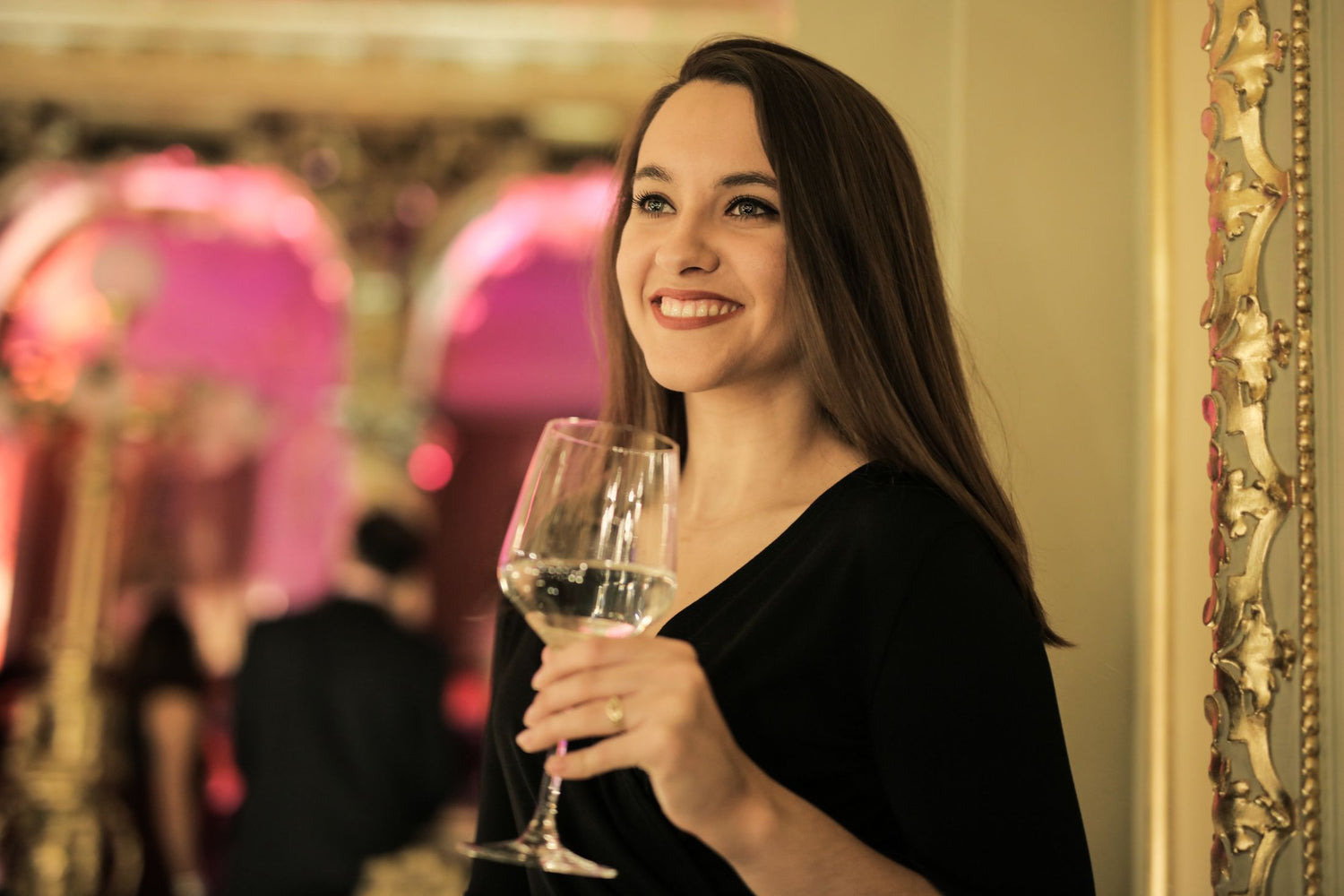Smiling woman in a black dress raising a glass of white wine at an elegant indoor celebration — wine gifts for her.