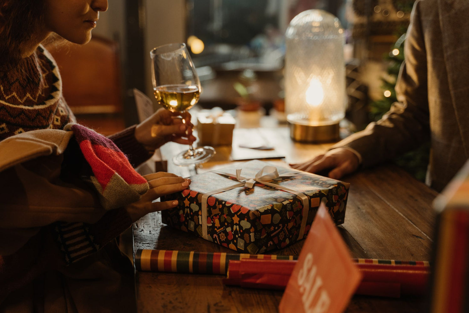 Two people sharing a wrapped gift at a festive Christmas table with wine glasses and a glowing lantern — wine gifts under £25.