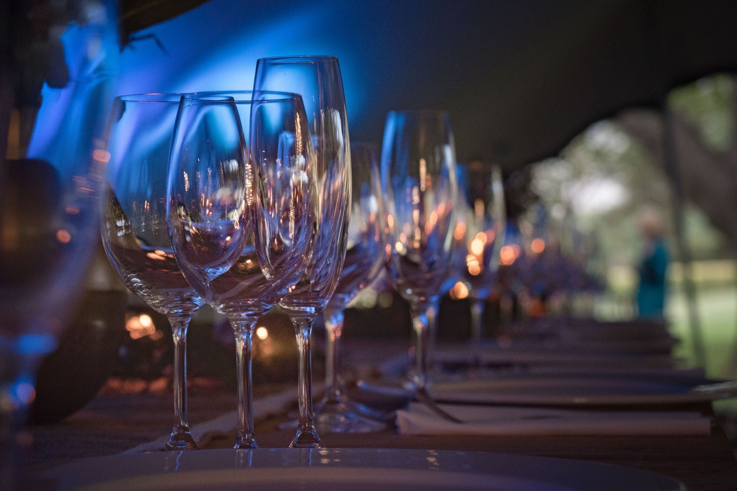 Rows of crystal wine glasses set along an elegantly dressed long dinner table under atmospheric blue lighting — great wines under £10-20.
