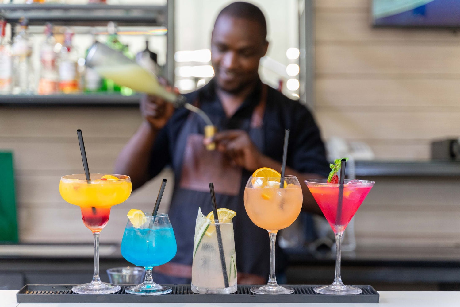 Bartender pouring a drink behind five colourful cocktails in various glasses at a modern bar