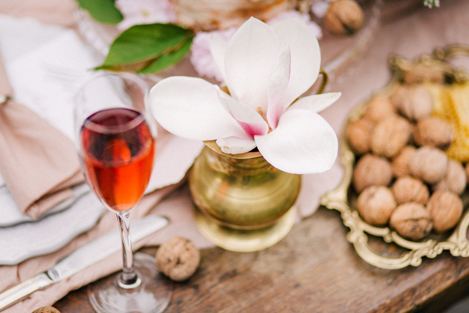 A glass of pink rosé wine beside a magnolia blossom in a brass vase, elegant tabletop styling for Pink Orchid