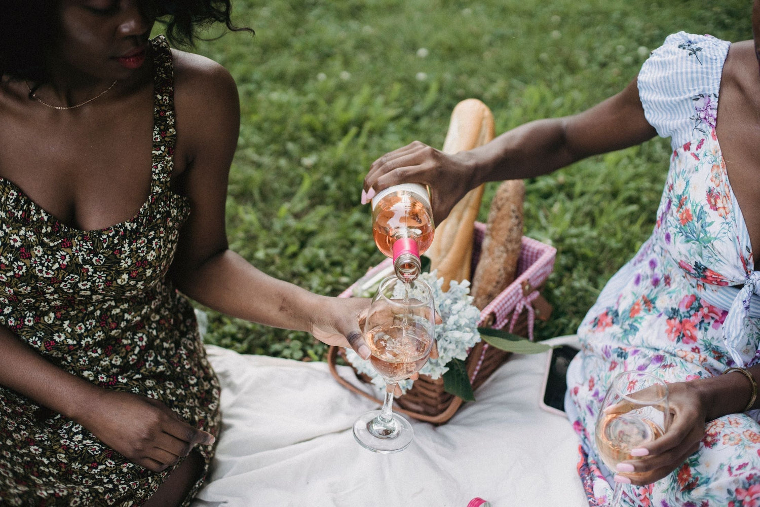 Friends pouring pale rosé wine on a sunny garden picnic, wicker basket and baguettes nearby, celebrating with rosé wine cases
