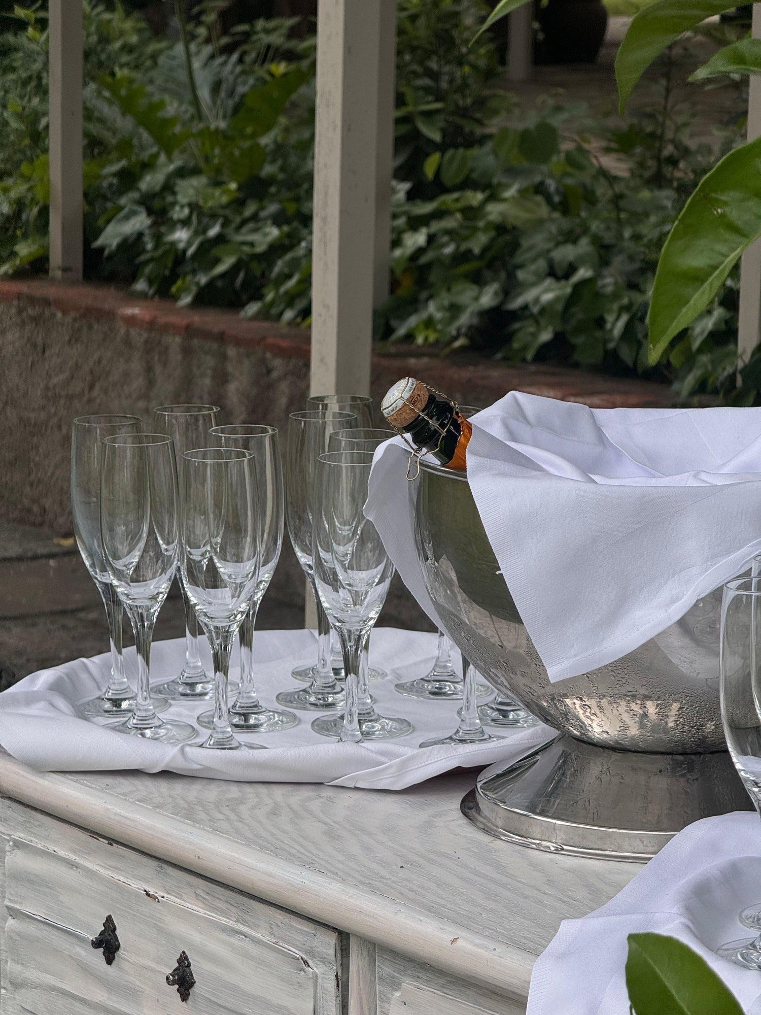 Multiple champagne flutes set out on white linen beside a chilled bottle in an ice bucket in a garden, sparkling wine celebration