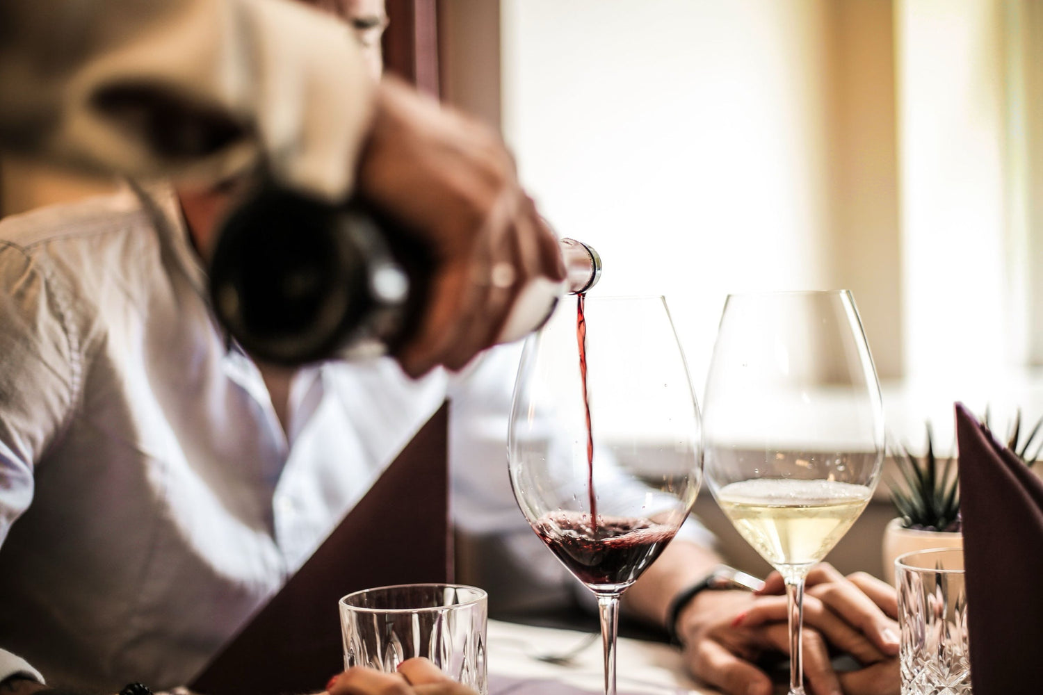 A waiter pouring red wine into a glass at a restaurant table, with white wine and menus alongside a dining couple