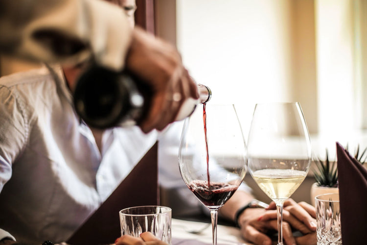 A waiter pouring red wine into a glass at a restaurant table, with white wine and menus alongside a dining couple