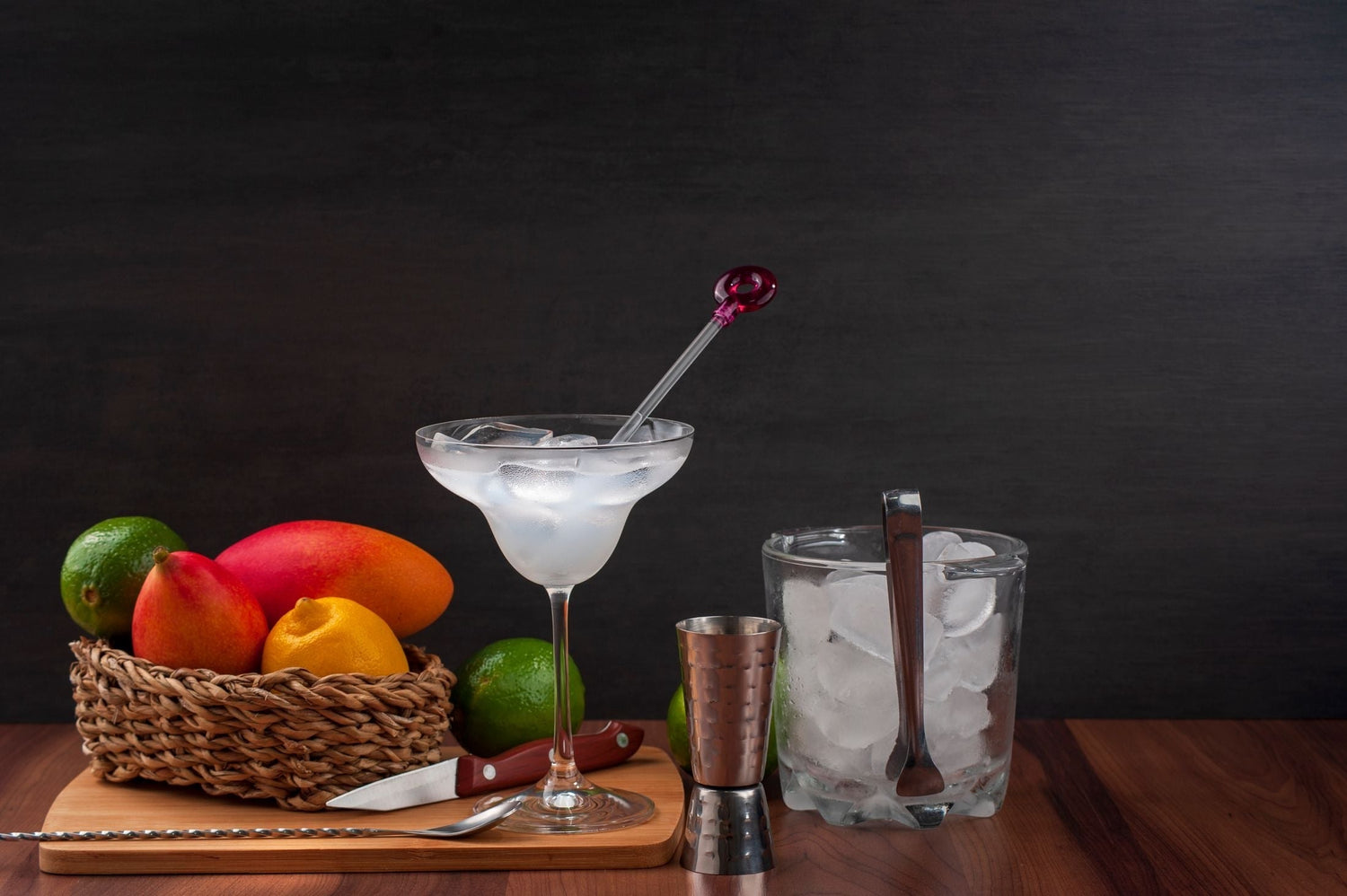 Margarita glass with ice beside a cocktail jigger, ice bucket, and a basket of fresh mangoes and limes — tequila cocktail ingredients