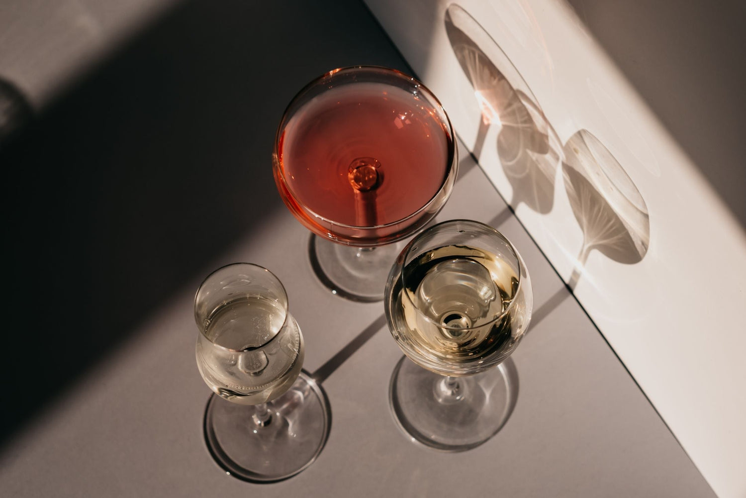 Overhead view of three wine glasses — a rose coupe, a white wine glass and a sparkling flute — casting dramatic shadows on a grey surface