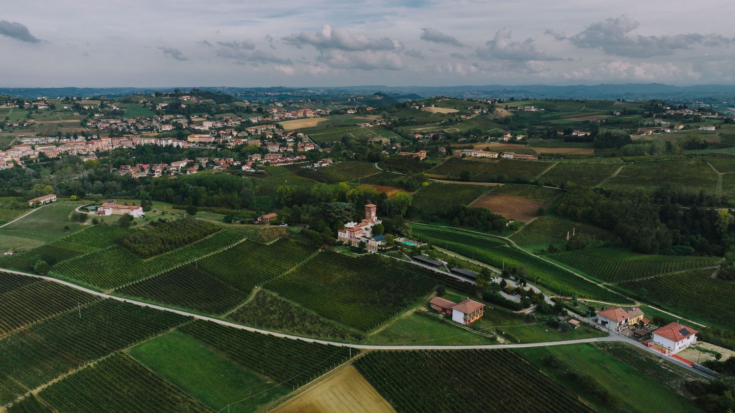 Aerial view of an Italian wine estate with a salmon-pink villa and tower surrounded by sweeping vineyard rows, rolling hills beyond