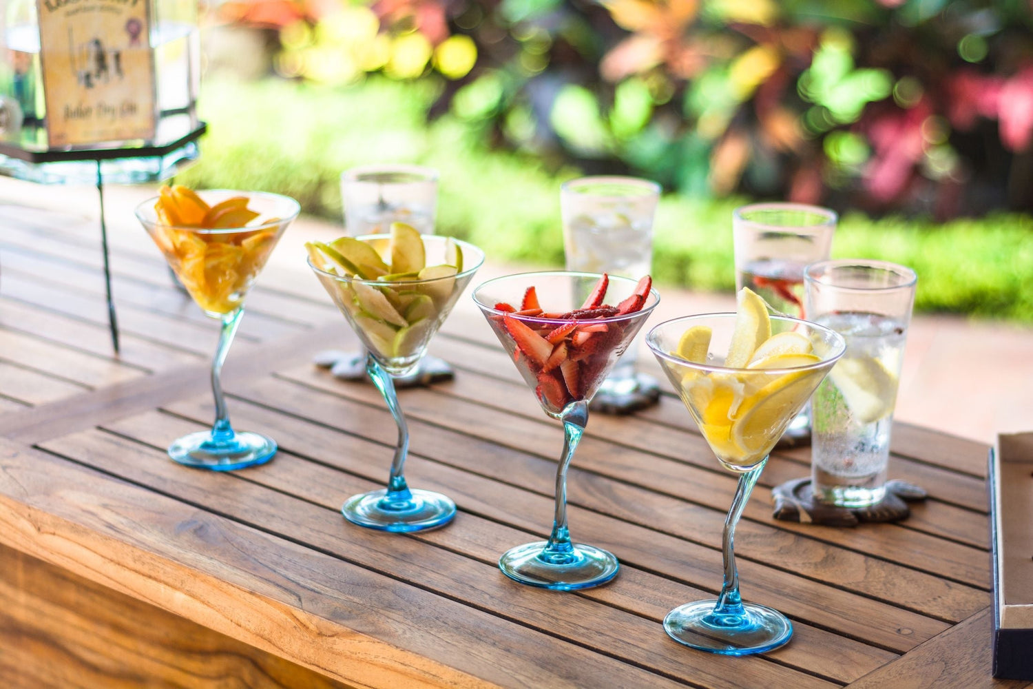 Colourful cocktail glasses filled with sliced fruit garnishes — orange, apple, strawberry and lemon — on a wooden outdoor table