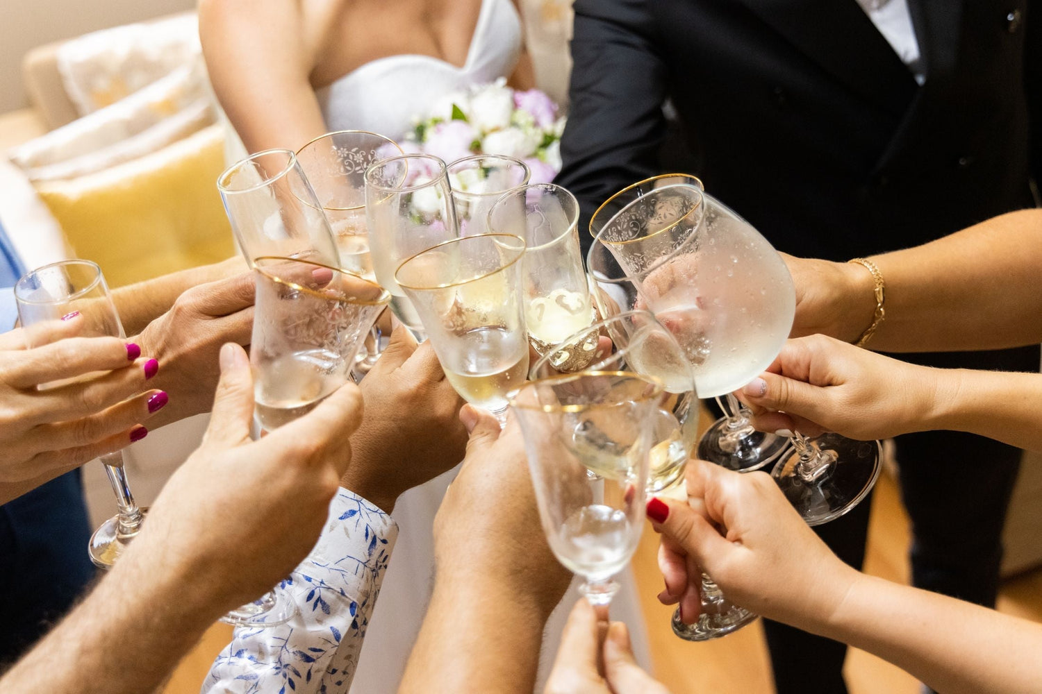 Hands raising champagne flutes in a celebratory toast at a wedding, bride in white dress with floral bouquet visible behind