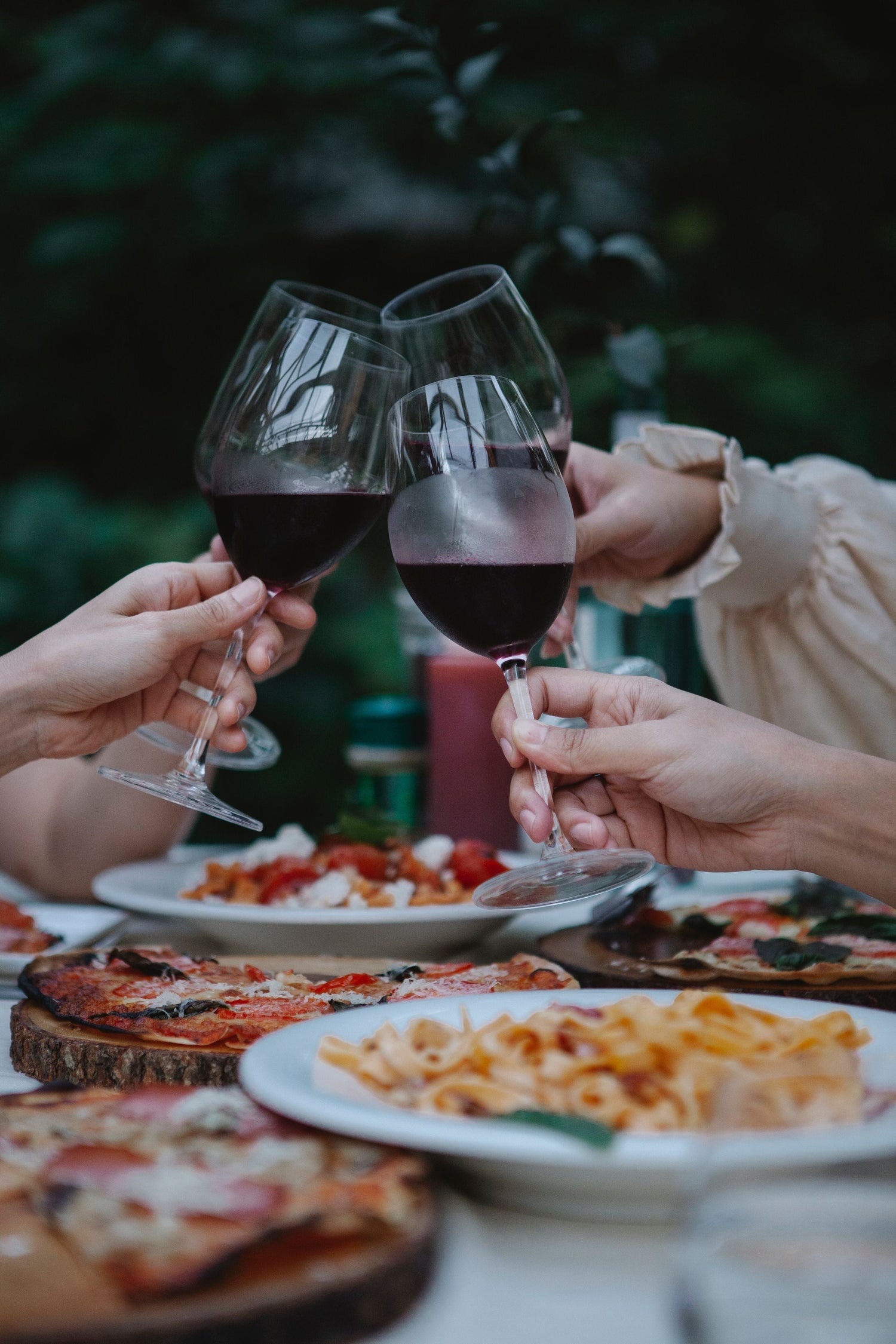 Two friends clink red wine glasses at an alfresco table laden with pizza and pasta, celebrating wines from around the world