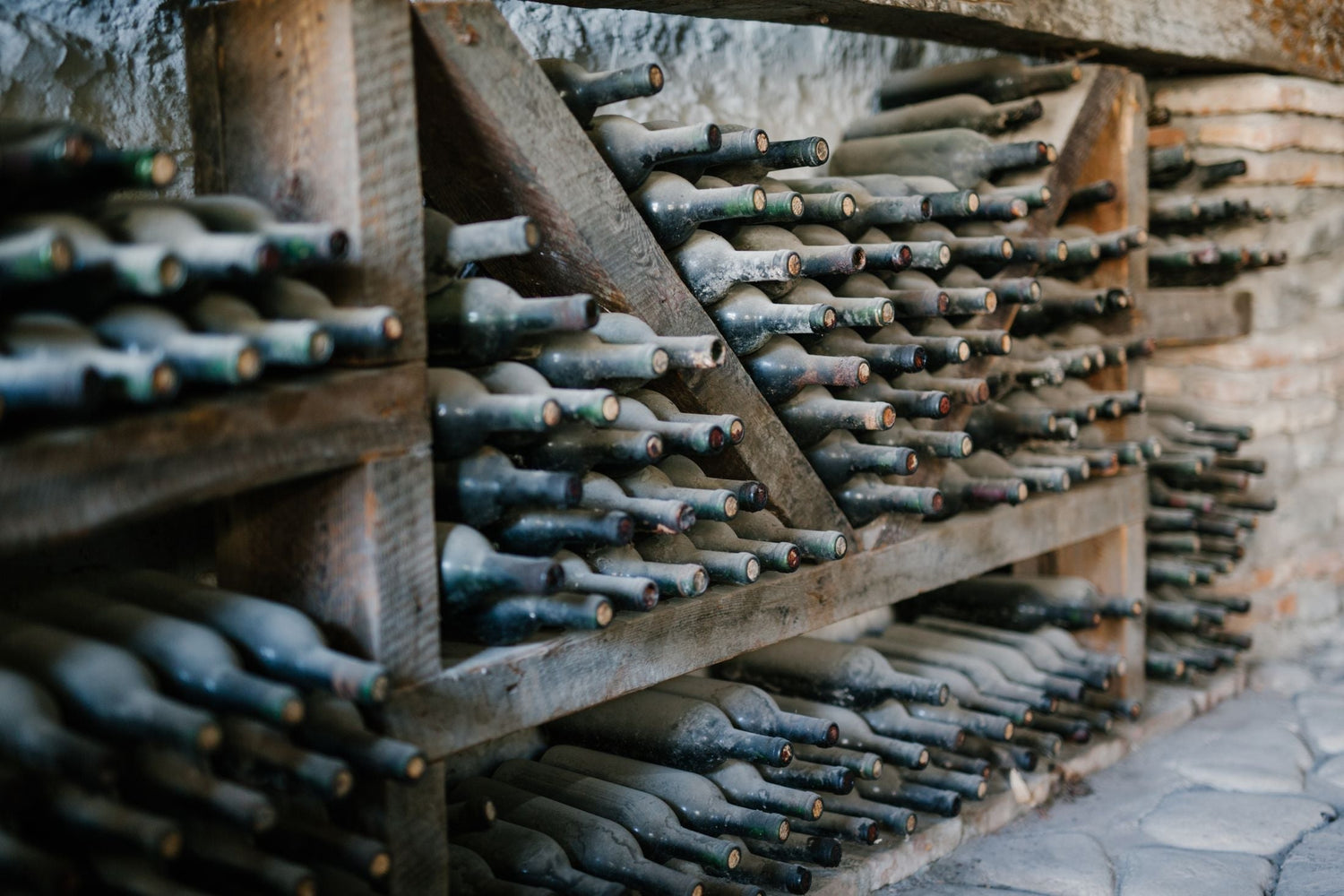 Dusty old wine bottles stored horizontally in a traditional angled wooden cellar rack, necks facing outward in a dimly lit stone cave