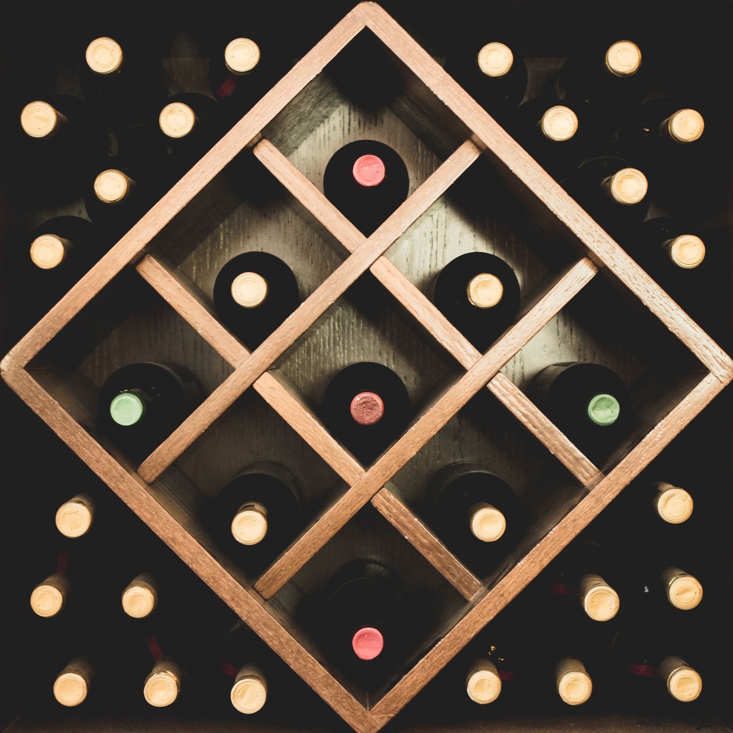 Wine bottles stored in a diamond-pattern wooden rack with coloured capsules visible — red, green and gold — set against dark wood