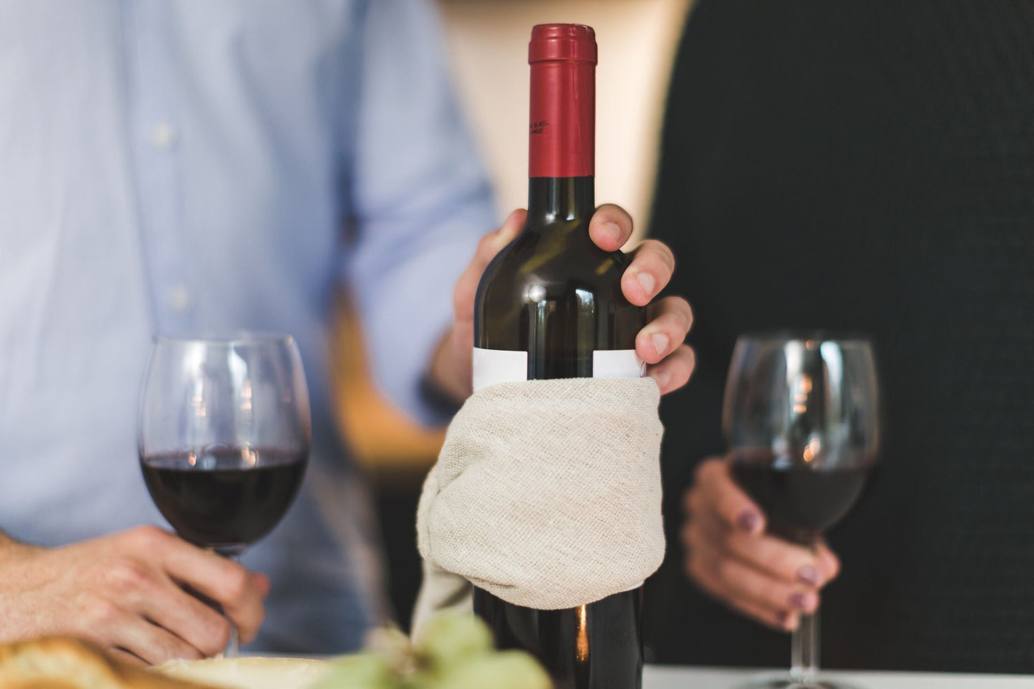A sommelier presents a red wine bottle wrapped in a cloth at a dining table where two guests hold their glasses ready to taste