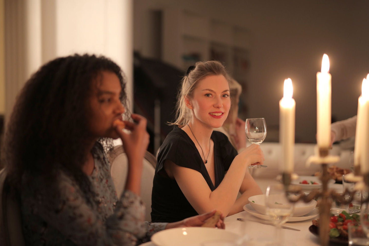 Two women enjoying a candlelit dinner together, one smiling and holding a wine glass, with a candelabra glowing beside them
