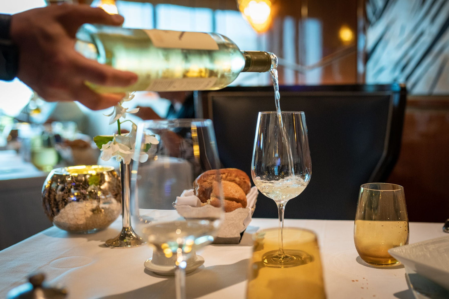 Sommelier pours white wine into a crystal glass at a fine dining table decorated with white flowers and a bread basket