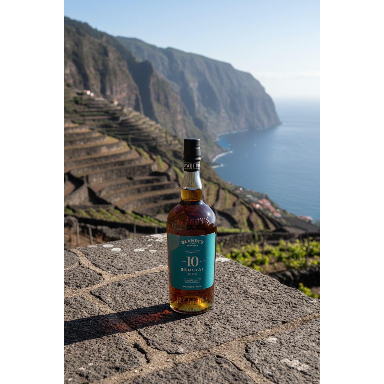 Blandy’s Sercial 10 Year Old Madeira wine bottle displayed on a stone terrace overlooking Madeira’s cliffside vineyards and the Atlantic Ocean.