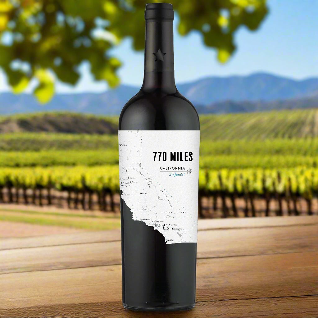 Place a bottle of 770 Miles Zinfandel on a wooden table with sunlit Californian vineyard rows in the background, highlighting the rugged landscape.