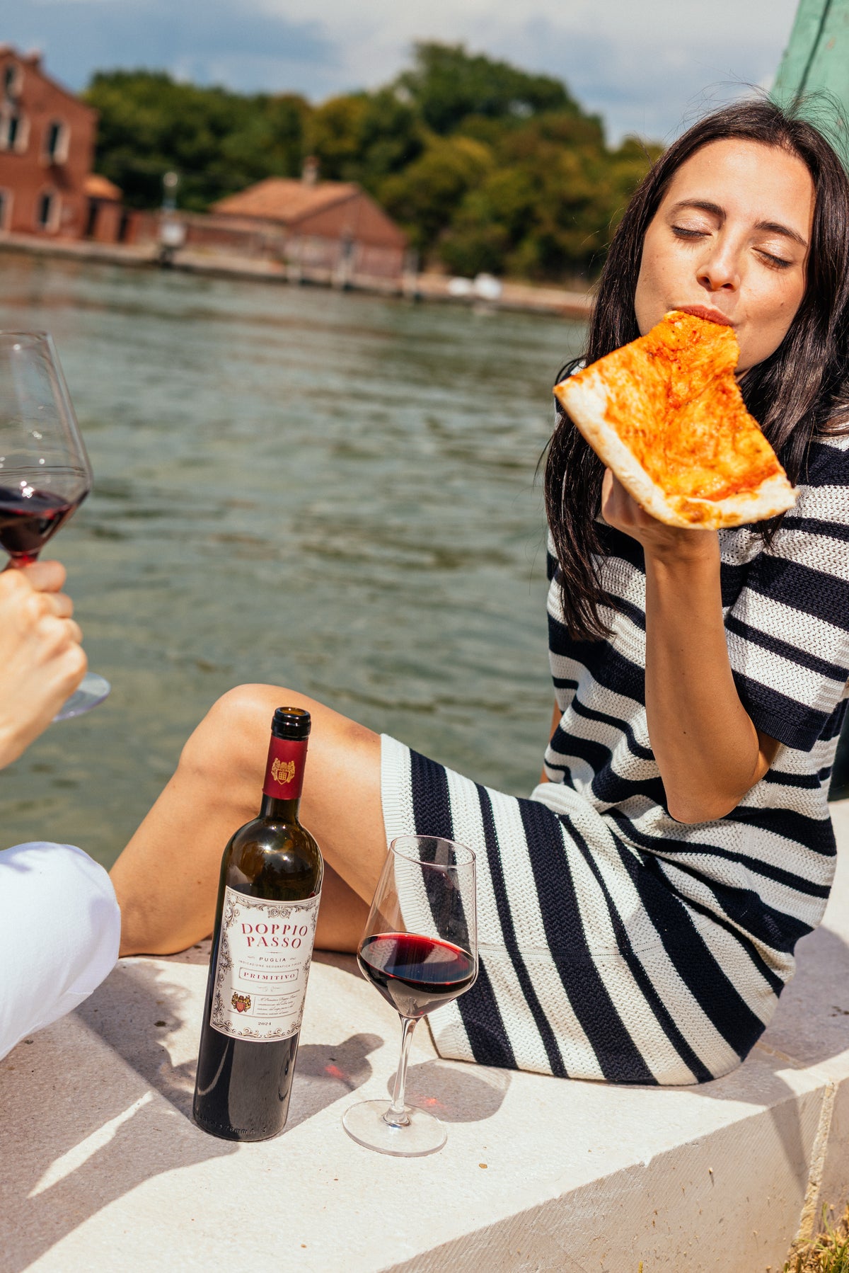 Woman eating pizza by a waterfront with a bottle of Doppio Passo Primitivo red wine and glass.