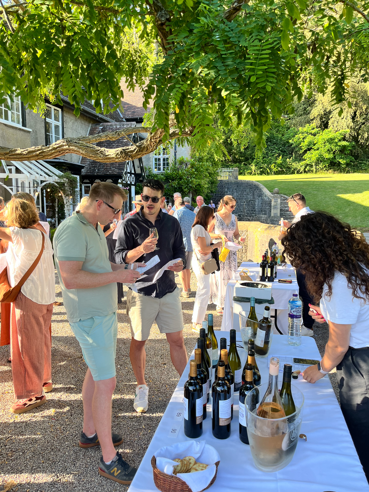A summer garden wine tasting hosted by The Wine Shop — guests tasting at tables under a tree